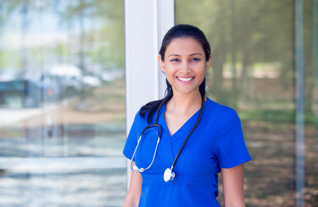 Closeup portrait of friendly, smiling confident female doctor, healthcare professional in blue scrubs with stethoscope, standing outside hospital background